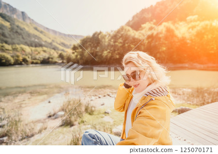 Woman, Mountains, River - A woman wearing a yellow jacket smiles as she sits on a wooden platform overlooking a mountain river. 125070171