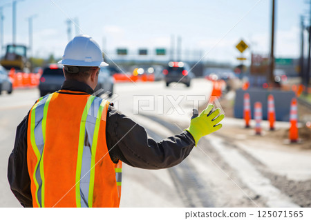 Construction worker wearing a hard hat and reflective vest is directing traffic on a busy road construction site 125071565