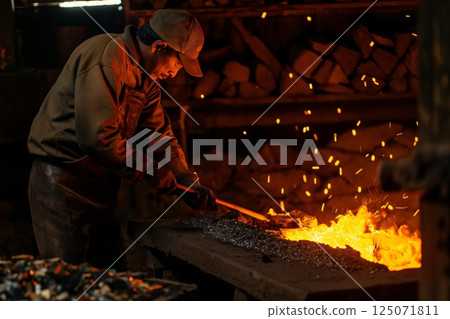 Blacksmith wearing protective leather apron and gloves, holding a hot metal rod with pliers in the forge 125071811