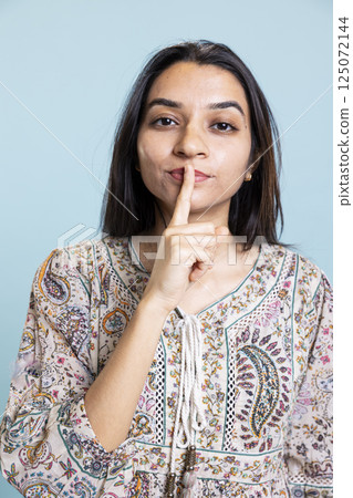 Indian young woman doing the hush mute symbol with finger over lips, female model asking for privacy and secrecy in studio. Silent mysterious person doing the shush gesture. 125072144