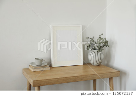 Beautiful Scandinavian interior. Green dry eucalyptus tree branches in vase on desk, table. Cup of coffee tea, old books. Empty wooden vertical picture frame mockup. Wall background. Elegant indoor. 125072157