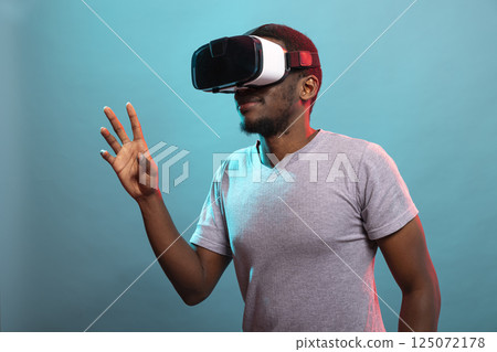 African american man raising his hand while experiencing an advanced VR simulation. Young black male individual with virtual reality glasses is immersed in a futuristic digital world. 125072178