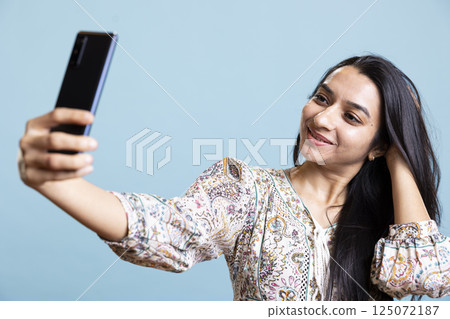 Pleased cheerful person using her smartphone to take selfies on social media. Jolly indian woman person taking photos using phone camera, standing against studio blue background. 125072187