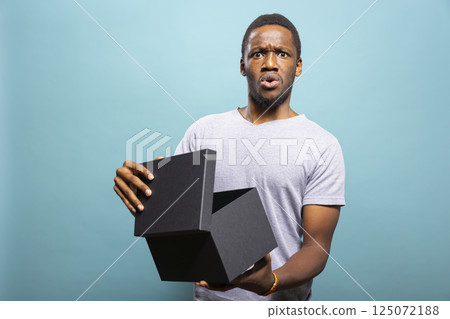 Amazed african american man opening a black giftbox, reacting with wide eyes in front of a blue background. Shocked individual unpacking a present, feeling amazed by the delivery surprise. Amazed african american man opening a black giftbox, reacting with wide eyes in front of a blue background. Shocked individual unpacking a present, feeling amazed by the delivery surprise. 125072188