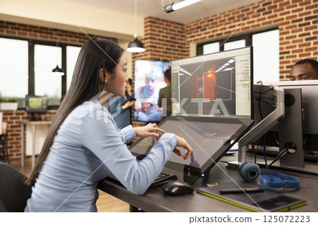Asian game developer creates professional content in the production department, using touchscreen display. Skilled woman focuses on developing 3D game scenes and environments, utilizing multi screens. Asian game developer creates professional content in the production department, using touchscreen display. Skilled woman focuses on developing 3D game scenes and environments, utilizing multi screens. 125072223