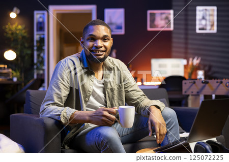 Black smiling guy sipping on fresh coffee in the morning before working, enjoying remote job from his cozy apartment. Young adult feeling happy and content with his career, multitasking. 125072225