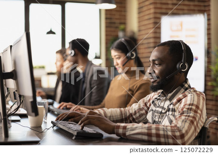 Black man in wheelchair and his multiracial coworkers seated at desks with computers and headsets, handling client calls. African American male customer service agent working in brick wall workspace. 125072229