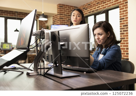 Selective focus on caucasian woman focused on computer screen, reviewing product designs in brick wall office. Female graphics designer analyzing images on desktop pc as asian manager stands nearby. Selective focus on caucasian woman focused on computer screen, reviewing product designs in brick wall office. Female graphics designer analyzing images on desktop pc as asian manager stands nearby. 125072236