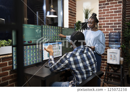 Black man and woman analyze market trends together on multiple screens in modern office with a brick wall. They focus on financial charts and stock data, working side by side through the night. Black man and woman analyze market trends together on multiple screens in modern office with a brick wall. They focus on financial charts and stock data, working side by side through the night. 125072238