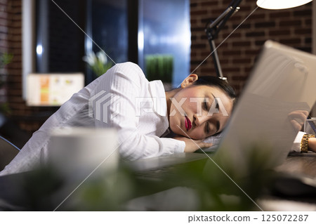 Overworked corporate manager sleeping at her office desk, taking a break during her night shift. Her laptop sits on the desk, highlighting the fatigue of long working hours in a modern workspace. Overworked corporate manager sleeping at her office desk, taking a break during her night shift. Her laptop sits on the desk, highlighting the fatigue of long working hours in a modern workspace. 125072287