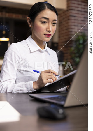Closeup of asian entrepreneur seated at her office desk, using a laptop and taking notes on a clipboard. The warm, dim lighting and contemporary designs highlights the night work environment. 125072289