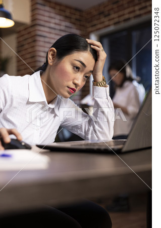 Asian female manager showing signs of fatigue while working late at her desk. Stressed businesswoman leans on one hand and looks at her laptop, as her blurred coworker reviews documents behind her. Asian female manager showing signs of fatigue while working late at her desk. Stressed businesswoman leans on one hand and looks at her laptop, as her blurred coworker reviews documents behind her. 125072348