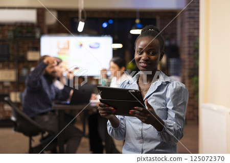 African american businesswoman holding her tablet and looking at camera in modern workspace. Portrait of black female financial advisor meeting deadlines, working late at night in startup office. African american businesswoman holding her tablet and looking at camera in modern workspace. Portrait of black female financial advisor meeting deadlines, working late at night in startup office. 125072370