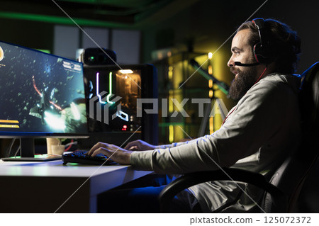 Upbeat man having fun with his friends playing videogames, chatting online using headphones microphone. Jolly gamer using computer with powerful graphics to enjoy multiplayer experience with teammates 125072372