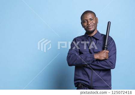 African american bodyguard posing with a baton and uniform in studio, showing confidence for his safeguarding duty. Skilled patrol officer standing against blue background on the job. 125072418