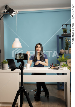 Beauty influencer sits at desk in a studio with a blue backdrop, engaging with her online audience during livestream. Perfect for themes of vlogging, content creation, and influencer marketing. 125072425