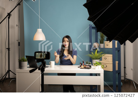 Young asian blogger recording a cosmetic review video in a studio with a camera on a tripod. She sits at a desk with makeup products, using professional lighting and a blue background. 125072427