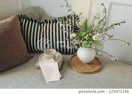Spring scandinavian living room interior. Blank greeting card invitation mockup, pink envelope. Sofa, linen striped, checkered cushions, cup of coffee. Cherry plum blossom tulips in vase. Elegant home Spring scandinavian living room interior. Blank greeting card invitation mockup, pink envelope. Sofa, linen striped, checkered cushions, cup of coffee. Cherry plum blossom tulips in vase. Elegant home 125075146