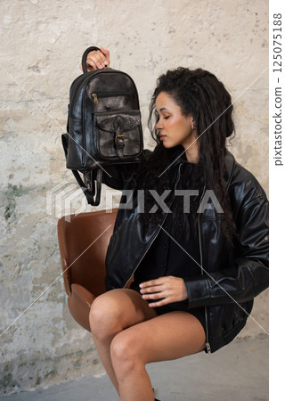 Indoors portrait of young beautiful fashionable woman wearing black leather jacket and short skirt posing with a backpack Indoors portrait of young beautiful fashionable woman wearing black leather jacket and short skirt posing with a backpack 125075188