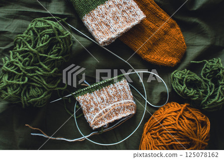 Dark green and brown yarn ball with knitted socks and knitting needles. Wool threads with deep green background. Woven crochet close up. Knitting hobby. Knitwear concept. Needlework background.  125078162