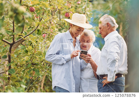 Woman showing something on the smartphone. Daughter is with her senior mother and father is in the garden Woman showing something on the smartphone. Daughter is with her senior mother and father is in the garden 125078876