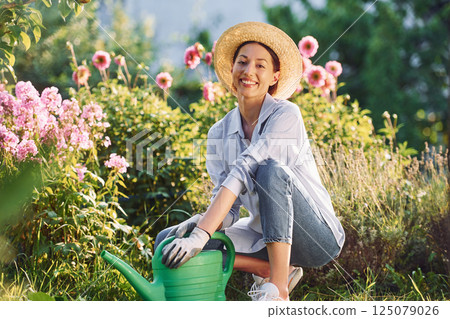 Preparing for watering the flowers. Young cheerful woman is in the garden at daytime Preparing for watering the flowers. Young cheerful woman is in the garden at daytime 125079026