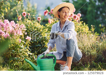 Preparing for watering the flowers. Young cheerful woman is in the garden at daytime 125079028