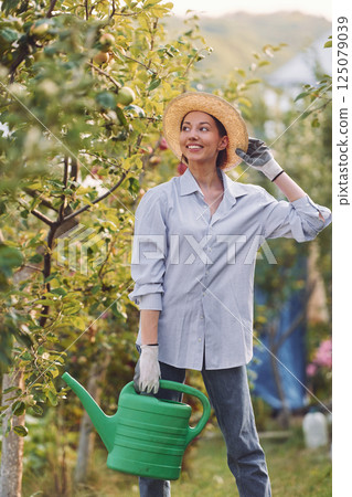 Standing and holding watering can. Young cheerful woman is in the garden at daytime Standing and holding watering can. Young cheerful woman is in the garden at daytime 125079039