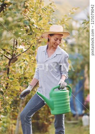 Standing and holding watering can. Young cheerful woman is in the garden at daytime Standing and holding watering can. Young cheerful woman is in the garden at daytime 125079042