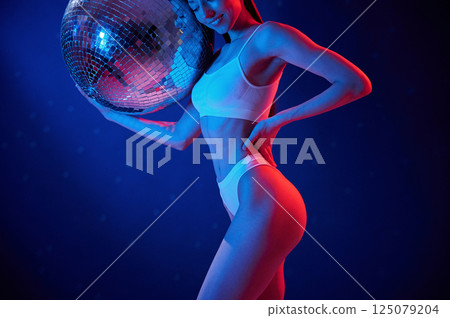 With shiny mirrored ball. Young woman in underwear is in the studio with neon lights With shiny mirrored ball. Young woman in underwear is in the studio with neon lights 125079204