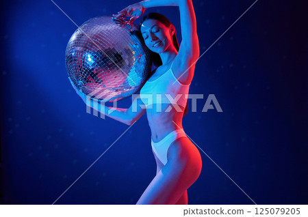 With shiny mirrored ball. Young woman in underwear is in the studio with neon lights With shiny mirrored ball. Young woman in underwear is in the studio with neon lights 125079205