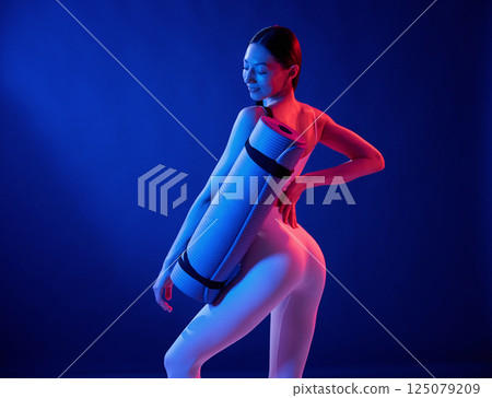 Standing with yoga mat. Young woman in sportive clothes is in the studio with neon lights Standing with yoga mat. Young woman in sportive clothes is in the studio with neon lights 125079209