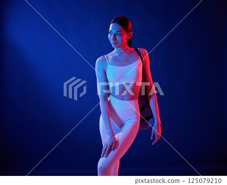Standing with yoga mat. Young woman in sportive clothes is in the studio with neon lights Standing with yoga mat. Young woman in sportive clothes is in the studio with neon lights 125079210