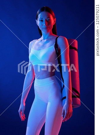 Standing with yoga mat. Young woman in sportive clothes is in the studio with neon lights 125079211
