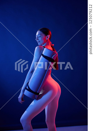 Standing with yoga mat. Young woman in sportive clothes is in the studio with neon lights Standing with yoga mat. Young woman in sportive clothes is in the studio with neon lights 125079212