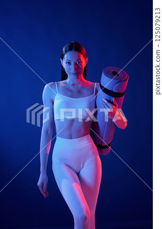 Front view. Holding the mat. Young woman in sportive clothes is in the studio with neon lights Front view. Holding the mat. Young woman in sportive clothes is in the studio with neon lights 125079213