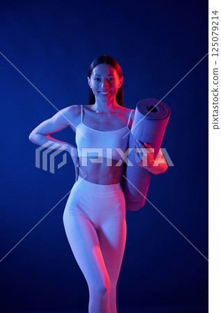 Front view. Holding the mat. Young woman in sportive clothes is in the studio with neon lights Front view. Holding the mat. Young woman in sportive clothes is in the studio with neon lights 125079214