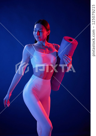 Front view. Holding the mat. Young woman in sportive clothes is in the studio with neon lights 125079215