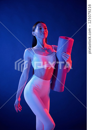 Front view. Holding the mat. Young woman in sportive clothes is in the studio with neon lights Front view. Holding the mat. Young woman in sportive clothes is in the studio with neon lights 125079216
