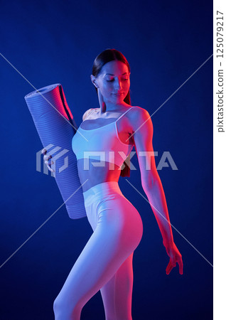Front view. Holding the mat. Young woman in sportive clothes is in the studio with neon lights 125079217