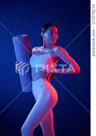 Ready for exercises. With yoga mat. Young woman in sportive clothes is in the studio with neon lights 125079218