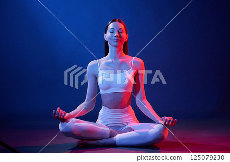 Lotus pose. Young woman in sportive clothes is in the studio with neon lights Lotus pose. Young woman in sportive clothes is in the studio with neon lights 125079230