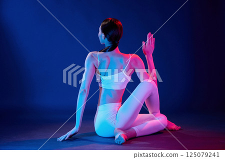 Rear view. Sitting and doing exercises. Young woman in sportive clothes is in the studio with neon lights Rear view. Sitting and doing exercises. Young woman in sportive clothes is in the studio with neon lights 125079241
