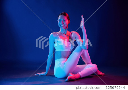 Sitting on the floor and doing exercises. Young woman in sportive clothes is in the studio with neon lights Sitting on the floor and doing exercises. Young woman in sportive clothes is in the studio with neon lights 125079244