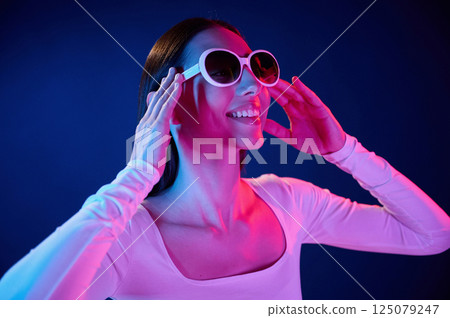 Smiling and wearing sunglasses. Portrait of young woman that is indoors in neon lighting Smiling and wearing sunglasses. Portrait of young woman that is indoors in neon lighting 125079247