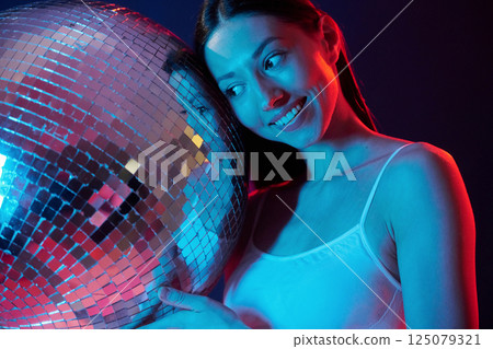 Party time. Holding mirrored ball. Young woman in underwear is in the studio with neon lights 125079321