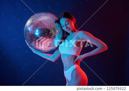 Party time. Holding mirrored ball. Young woman in underwear is in the studio with neon lights 125079322