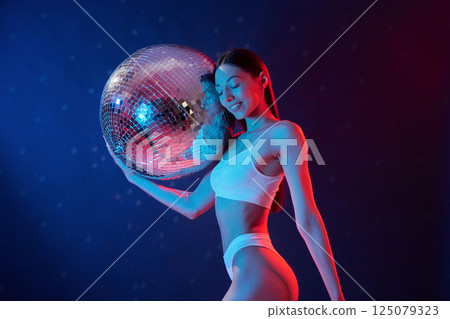 Party time. Holding mirrored ball. Young woman in underwear is in the studio with neon lights Party time. Holding mirrored ball. Young woman in underwear is in the studio with neon lights 125079323