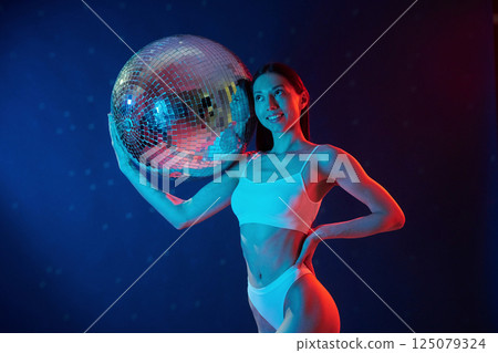 Party time. Holding mirrored ball. Young woman in underwear is in the studio with neon lights 125079324