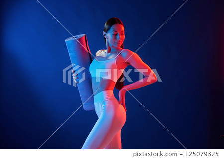 Young woman in sportive clothes is in the studio with neon lights. Holding mat Young woman in sportive clothes is in the studio with neon lights. Holding mat 125079325